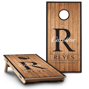 May include: Two rectangular cornhole boards featuring a faux wood plank pattern. Each board has a circular hole at the top, a large black letter R, the names Caroline or Henry, the surname REYES, and the text ESTABLISHED JUNE 2027 in black.