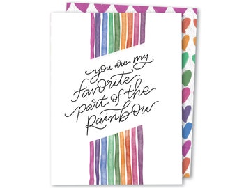 May include: A greeting card with a white background and the text "you are my favorite part of the rainbow" in black script. Vertical watercolor stripes in rainbow colors frame the text. The card's edge features a pattern of colorful hearts.