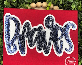 May include: A red t-shirt featuring the word Braves in dark blue and light blue sequins with a white outline. The text is written in a cursive script font. A small white circular logo with the text Carrie Made Mine is in the bottom right corner.