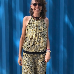 Könnte beinhalten: Eine Frau trägt einen gelben und schwarzen Tierdruck-Jumpsuit mit einem schwarzen Halfter-Oberteil und einer Perlenkette. Sie steht vor einer blauen Wand und trägt Sandalen.