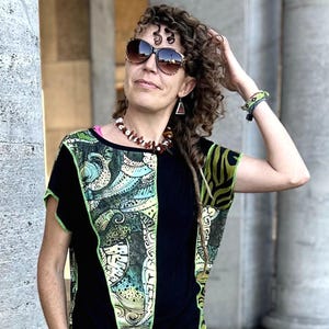 Könnte beinhalten: Eine Frau trägt ein schwarzes Oberteil mit einem bunten Muster und grünen Verzierungen, kombiniert mit einer leuchtend grünen Hose. Sie trägt eine Sonnenbrille, eine Halskette und ein Armband. Der Hintergrund zeigt große Steinsäulen.