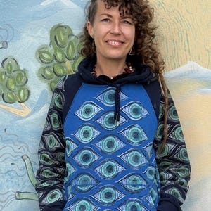 Puede incluir: Una sudadera con capucha azul con un patrón de ojos repetido en tonos azules y turquesas. Las mangas tienen un diseño estampado y la capucha es azul marino. La persona está de pie frente a un mural con un mono y una escena submarina.