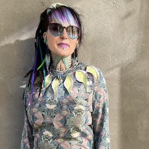 Puede incluir: Una sudadera de manga larga con un estampado floral en tonos grises, rosas y azules. La sudadera tiene un cuello alto con detalles en forma de hojas amarillas y verdes. La persona lleva pantalones cortos vaqueros azules claros y leggings morados.