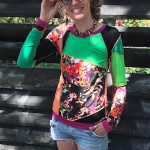 Könnte beinhalten: Eine Frau trägt ein farbenfrohes Langarmhemd mit einem Blumenmuster und einem geometrischen Design in Grün, Schwarz und Pink. Sie trägt eine blaue Jeans-Shorts und eine Sonnenbrille mit roten Rahmen.