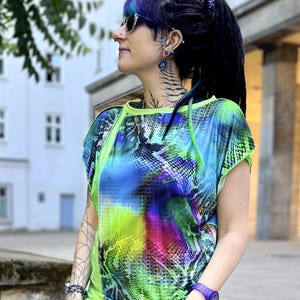 Op de afbeelding: Een felgekleurde top met korte mouwen en een kleurrijk slangenpatroon in blauw, groen en roze. De top heeft neon groene randen en wordt gecombineerd met een bijpassende groene broek. Een paars horloge en sieraden maken de look compleet.