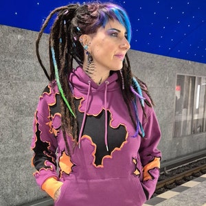 Op de afbeelding: Een paarse hoodie met zwarte en oranje wolkachtige ontwerpen. De hoodie heeft een capuchon met trekkoord en een kangoeroezak. De persoon die de hoodie draagt heeft blauw en paars haar.
