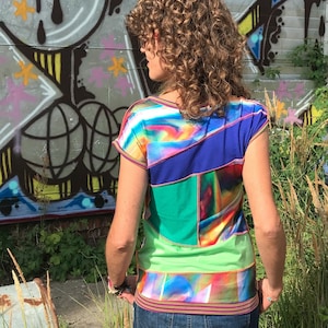 Könnte beinhalten: Ein kurzärmliges Shirt mit Rundhalsausschnitt und einem bunten, abstrakten Druck. Das Shirt besteht aus einem Patchwork aus verschiedenen Stoffen in Blautönen, Grün, Rosa und Weiß.