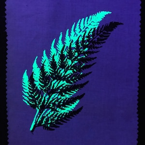 Op de afbeelding: Een zwarte varenblad gedrukt op een paarse stoffen achtergrond. De varen is omrand met een heldergroene kleur.