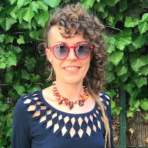 Könnte beinhalten: Eine Frau mit lockigem Haar trägt eine rote Sonnenbrille und eine Halskette aus Bernstein. Sie trägt ein dunkelblaues Shirt mit einem Ausschnitt.