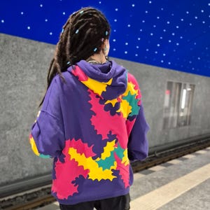 May include: A purple hoodie with a colourful abstract design featuring pink, yellow, and green shapes. The hoodie has a drawstring hood and a kangaroo pocket.