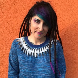 Puede incluir: Una mujer con el pelo morado y azul lleva un suéter azul y blanco con un cuello blanco recortado.