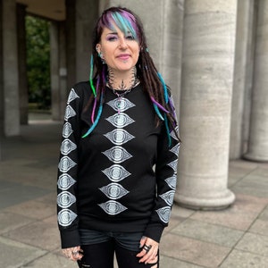 May include: A black long-sleeved shirt with a white eye pattern. The shirt has a crew neck and long sleeves. The model is wearing the shirt with black trousers and has colourful dreadlocks.