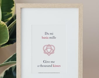 May include: Framed print with a light wood grain frame. The print features the text "Da mi basia mille" and "Give me a thousand kisses" in red and black. A red heart-shaped knot design is centered between the text.