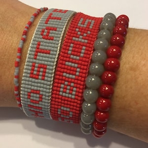 May include: A collection of bracelets in shades of red and gray. One bracelet features the word "LOVE" in red against a gray background. Other bracelets include beaded and round stone designs. The bracelets are stacked on a wrist.