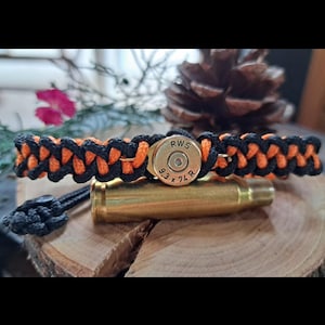 Könnte beinhalten: Ein geflochtenes Armband aus schwarzer und orangefarbener Kordel mit einer Messinghülse als zentralem Element. Die Hülse ist mit dem Text RWS und 9.3 x 74 R geprägt. Das Armband hat an einem Ende einen Verschluss aus einem schwarzen Knoten.