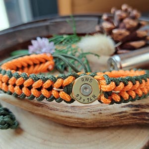 Peut inclure: Bracelet tressé avec un cordon orange et vert olive. Le bracelet comporte un étui de balle en laiton avec le texte "RWS 9.3 x 74 R". Le bracelet est présenté sur une surface en bois avec d'autres éléments naturels.