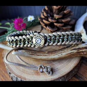 Peut inclure: Un bracelet en paracorde tressée avec une douille en laiton au centre portant l'inscription Hornady 6.5x62. Le bracelet est tissé avec des cordons beige, vert foncé et noir, et comprend un cordon de serrage beige avec un nœud à l'extrémité.