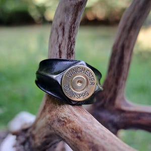 Pode incluir: Um anel preto com uma caixa de bala de ouro Winchester 3006 SPRG. O anel está em um chifre de veado marrom.