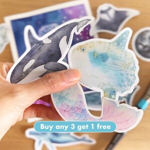Op de afbeelding: Een set kleurrijke aquarel stickers met verschillende zeedieren, waaronder een walvis, een zeemeermin, een maanvis en een mantarog. De stickers zijn gedrukt op wit, glanzend papier en hebben een matte afwerking. De tekst "Koop er 3, krijg er 1 gratis" is gedrukt op een wit banner.