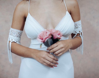 May include: A white wedding dress with a plunging neckline and lace detailing on the sleeves. The dress is paired with a bouquet of pink roses.