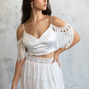 May include: A white satin crop top with crochet and fringe detailing on the shoulders, paired with a white tulle skirt with a crochet waistband.