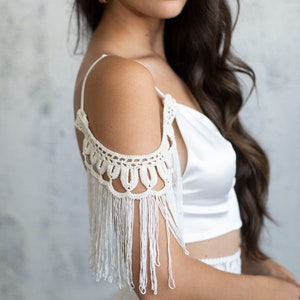 May include: A white satin crop top with a crocheted off-the-shoulder detail and fringe trim.