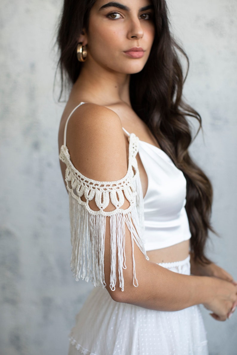 May include: A white satin crop top with a crocheted off-the-shoulder detail and fringe trim.
