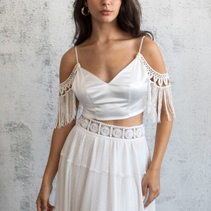 May include: A white satin crop top with crochet fringe detailing on the shoulders and a white crochet waistband. The top is paired with a white tiered skirt with a dotted pattern.