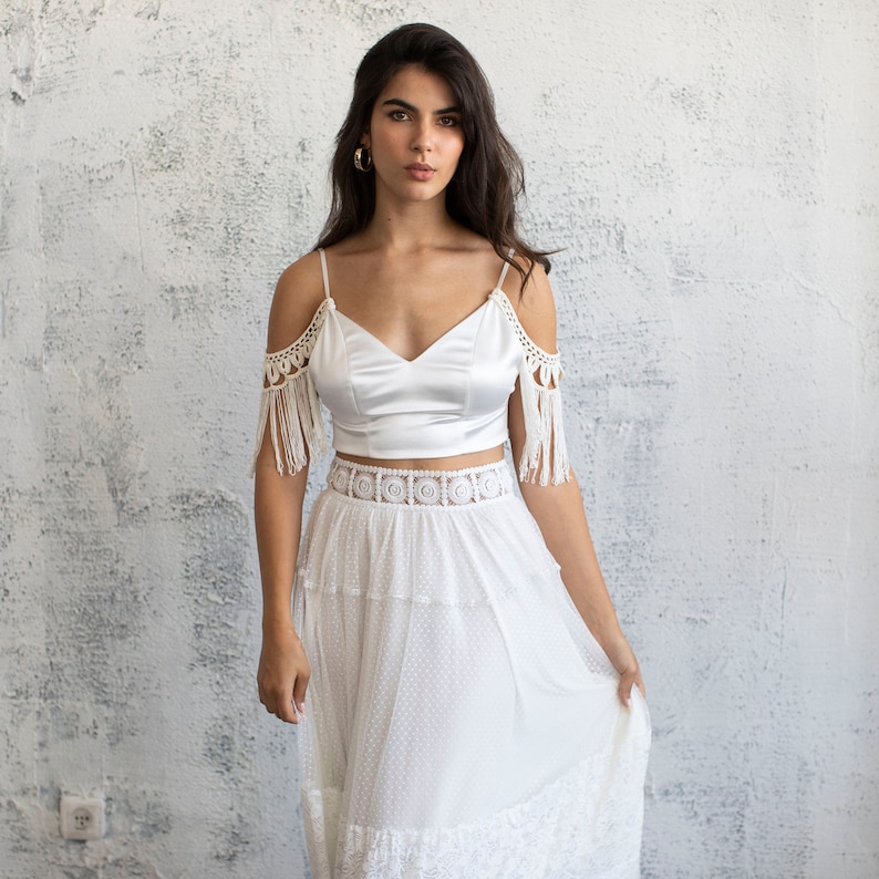 May include: A white satin crop top with crochet trim and fringe detailing. The top is paired with a white tulle skirt with a lace waistband and a dotted pattern.