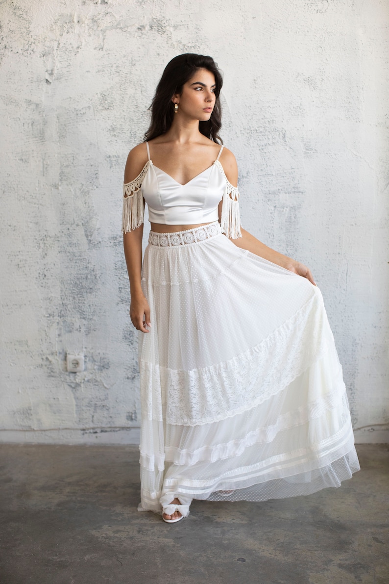 May include: A white bridal top with crochet fringe detailing on the shoulders and a white tulle skirt with lace trim. The skirt has multiple layers and a delicate polka dot pattern.