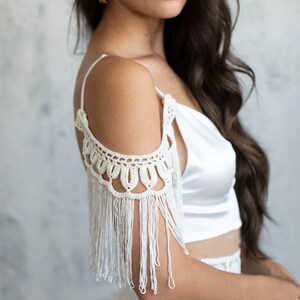 May include: A white satin crop top with a crocheted off-the-shoulder detail and fringe trim.