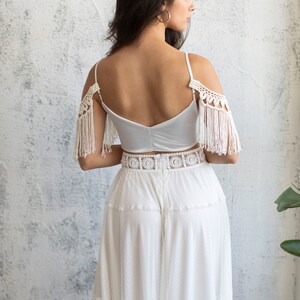 May include: A white crochet top with fringe detailing on the shoulders and a white lace skirt. The top has a low back and the skirt has a tiered design.