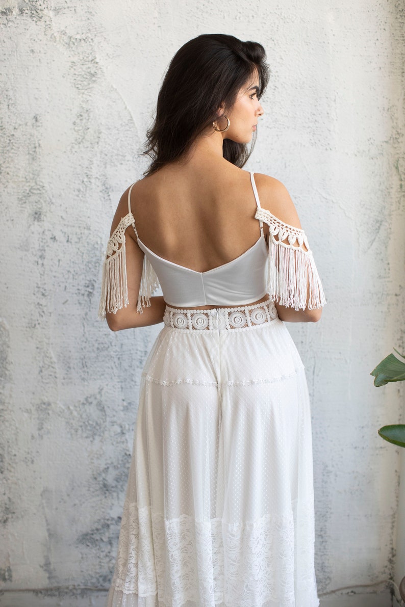 May include: A white crochet top with fringe detailing on the shoulders and a white lace skirt. The top has a low back and the skirt has a tiered design.