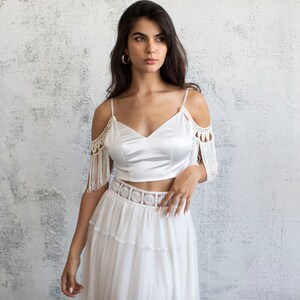 May include: A white off-the-shoulder crop top with crochet detailing and fringe. The top is paired with a white skirt with a crochet waistband and a tiered design.