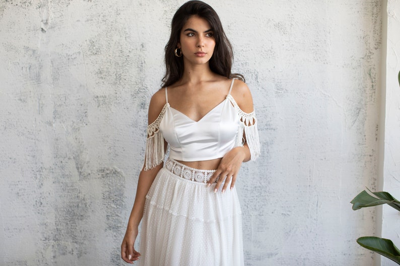 May include: A white off-the-shoulder crop top with crochet detailing and fringe. The top is paired with a white skirt with a crochet waistband and a tiered design.
