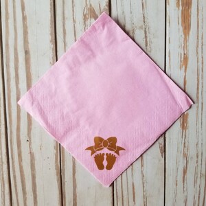 Peut inclure: Serviette en papier rose avec un motif en feuille d'or d'un n&oelig;ud et d'empreintes de pieds de b&eacute;b&eacute;.