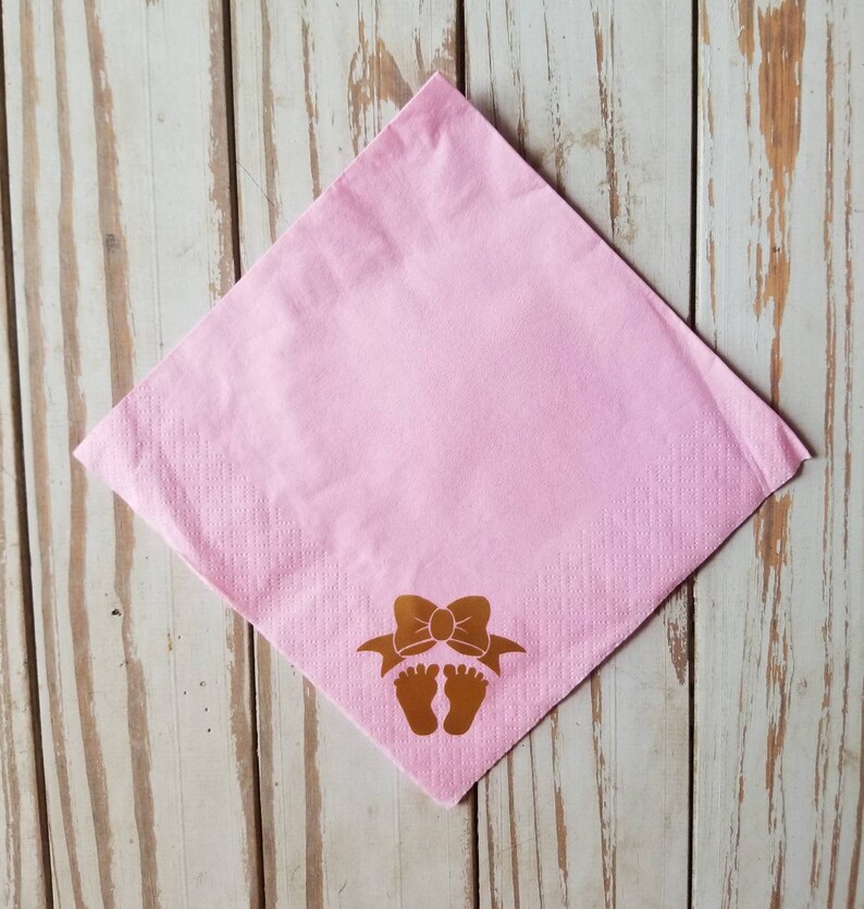 Peut inclure: Serviette en papier rose avec un motif en feuille d'or d'un n&oelig;ud et d'empreintes de pieds de b&eacute;b&eacute;.