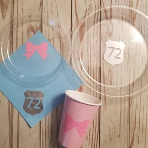 May include: Two clear plastic plates with a pink bow and a silver shield with the number 72 on a blue napkin. A pink cup with a pink bow and the number 72 on a silver shield.