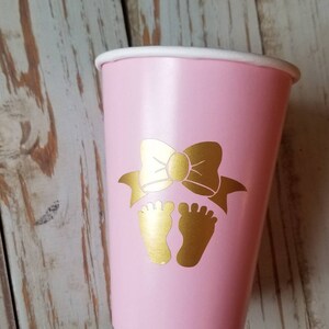 Peut inclure: Gobelet en papier rose avec un n&oelig;ud dor&eacute; et deux empreintes de pieds dor&eacute;es. Le gobelet est parfait pour une f&ecirc;te pr&eacute;natale.