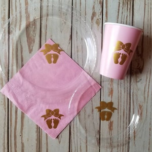 Peut inclure: Assiettes et gobelets de f&ecirc;te en plastique rose et transparent avec des empreintes de pieds de b&eacute;b&eacute; dor&eacute;es et des n&oelig;uds. Les assiettes sont transparentes avec une serviette rose sur le dessus.