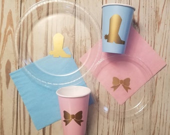 May include: A set of party supplies featuring a blue cup with a gold boot design, a pink cup with a gold bow design, a clear plastic plate with a gold bow design, and a clear plastic plate with a gold boot design. The plates and cups are on a light wood surface with blue and pink napkins.