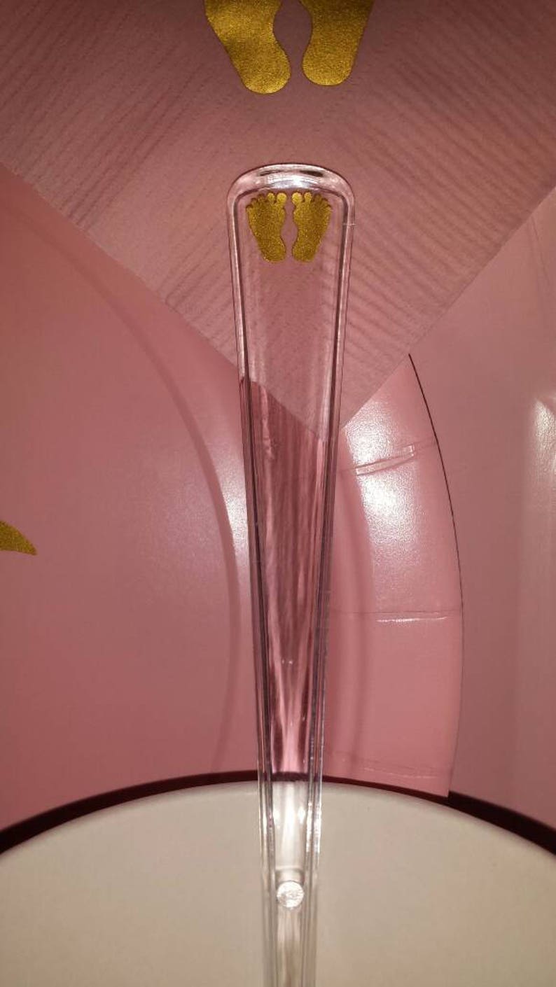 Peut inclure: Un serveur &agrave; g&acirc;teau en plastique transparent avec un motif de petite empreinte de pied dor&eacute;e. Le serveur est sur une surface rose.