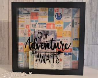 May include: Black framed shadow box with a travel-themed background of colorful vintage tickets. The box contains banknotes and the words "Our Adventure Awaits" in black script with an arrow. A decorative piece for saving money.