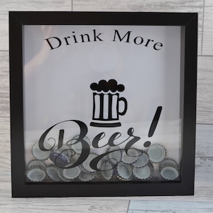 May include: Black framed shadow box with the words "Drink More Beer!" and a beer mug graphic. The box is filled with bottle caps. The background is a light wood grain.