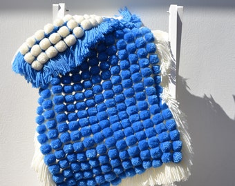 May include: A handmade blue and white pom-pom blanket. The blanket features a grid of blue pom-poms, white pom-poms, and white fringe. The blanket is draped over a white ladder. The blanket is a cozy home decor item.