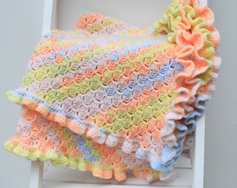 May include: A handmade, pastel-colored crochet blanket with a ruffled edge. The blanket features a mix of soft colors including peach, yellow, blue, and pink. The blanket is folded over a white shelf, showcasing the texture and design.