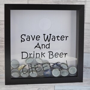 May include: A black framed shadow box with the text "Save Water And Drink Beer" and "Cheers" in black lettering. The box is filled with beer bottle caps. A white circle is at the top.