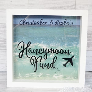 Pode incluir: Caixa de sombra com moldura branca com fundo de cena de praia. O texto "Christopher & Sasha's" está no topo, com "Honeymoon Fund" em itálico abaixo. Um avião preto está presente.