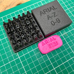 May include: A black Arial font stamp set with letters A-Z and numbers 0-9. The set includes a tray of individual stamps, a storage box with "ARIAL A-Z 0-9" text, and a pink stamp with "ARIAL 6MM" text. The items are on a green grid surface.