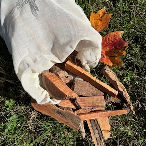 May include: A beige muslin bag with the text "FAIRWOOD FIRESTEELS" and a hand-and-dagger logo, spilling out pieces of reddish-brown wood and bark onto green grass. Two colorful autumn leaves are next to the bag.